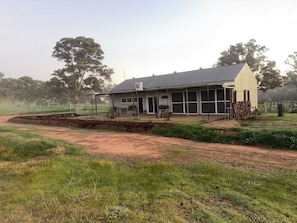 A single-story building with a large open porch area, surrounded by a fenced yard with a dirt path leading to the entrance.