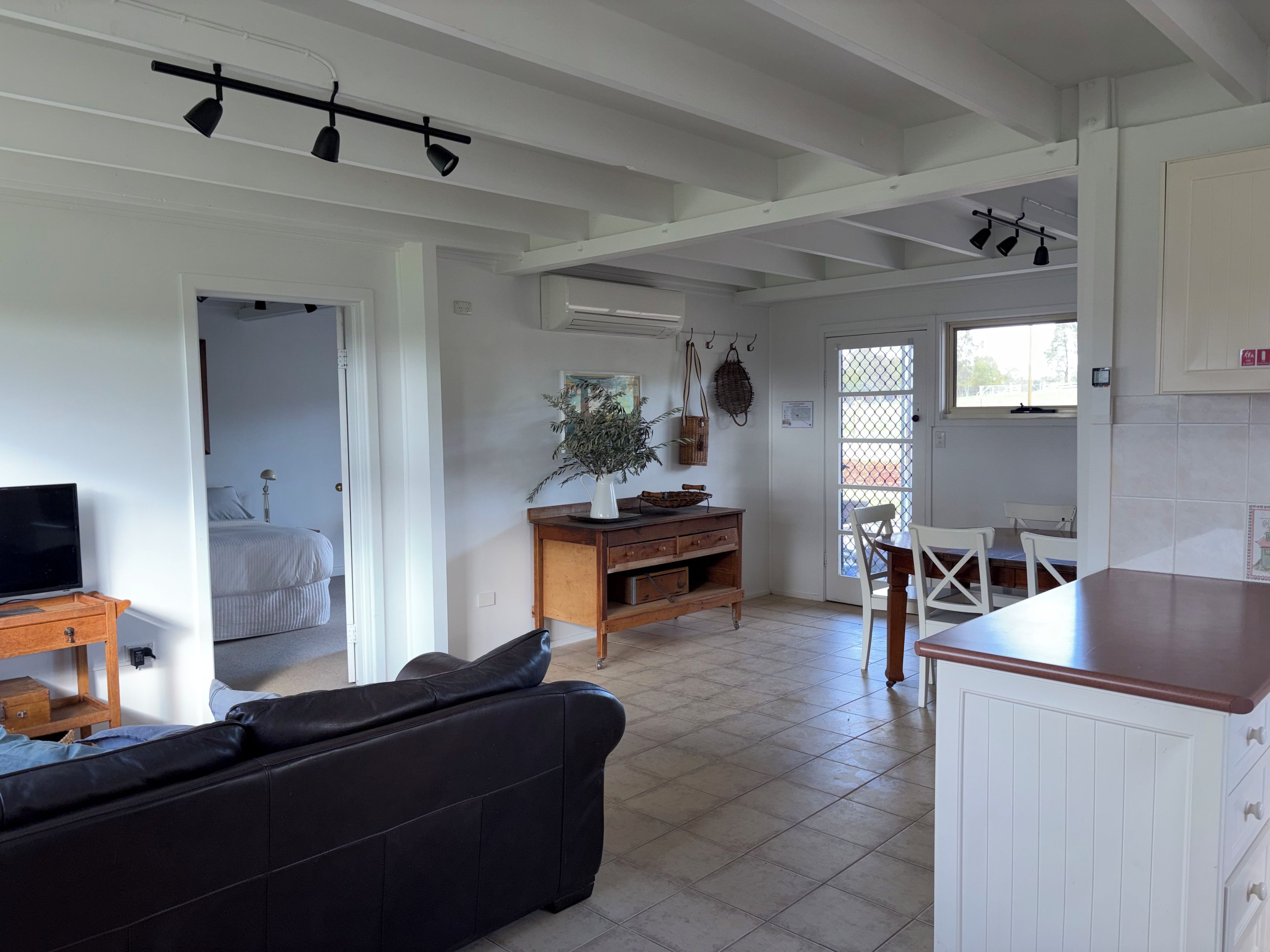 A room with a black leather sofa, a wooden dining table with chairs, a kitchen area with cabinets, and a door leading to another room.