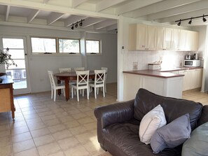 A spacious living area with a dining table, white chairs, a brown leather sofa, and a kitchen with white cabinets and appliances.