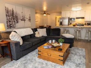 Ein Wohnzimmer mit einer Eckcouch, einer Holzkiste und einem Couchtisch. Es gibt eine Küchenzeile mit weißen Schränken und einem Kühlschrank.