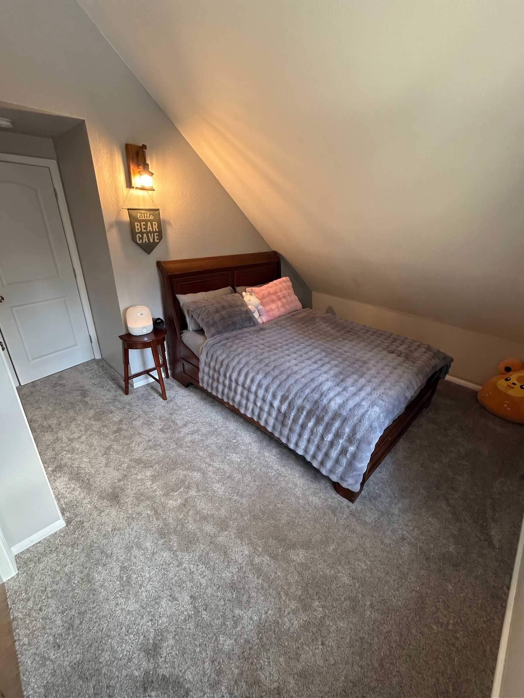 A bedroom with a sloped ceiling, a bed with a grey blanket, a bedside table, a lamp, and a wall-mounted sign.