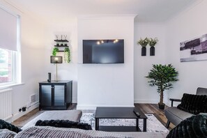 A modern living room with a flat-screen TV, a cabinet, a coffee table, a sofa, and a potted plant. - Stylish 3-Bed Home in Leigh | Garden & Parking (Greater Manchester)