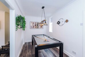 Ein Billardtisch mit Billardkugeln, eine hängende Leuchte, eine Uhr und ein Grünpflanzen an der Wand.