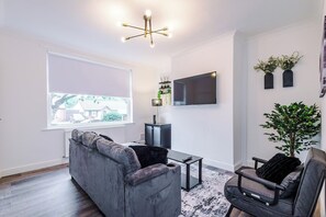 A modern living room with a grey sofa, a black armchair, a coffee table, a flat-screen TV mounted on the wall, and a large window with a view of a house outside. - Stylish 3-Bed Home in Leigh | Garden & Parking (Greater Manchester)