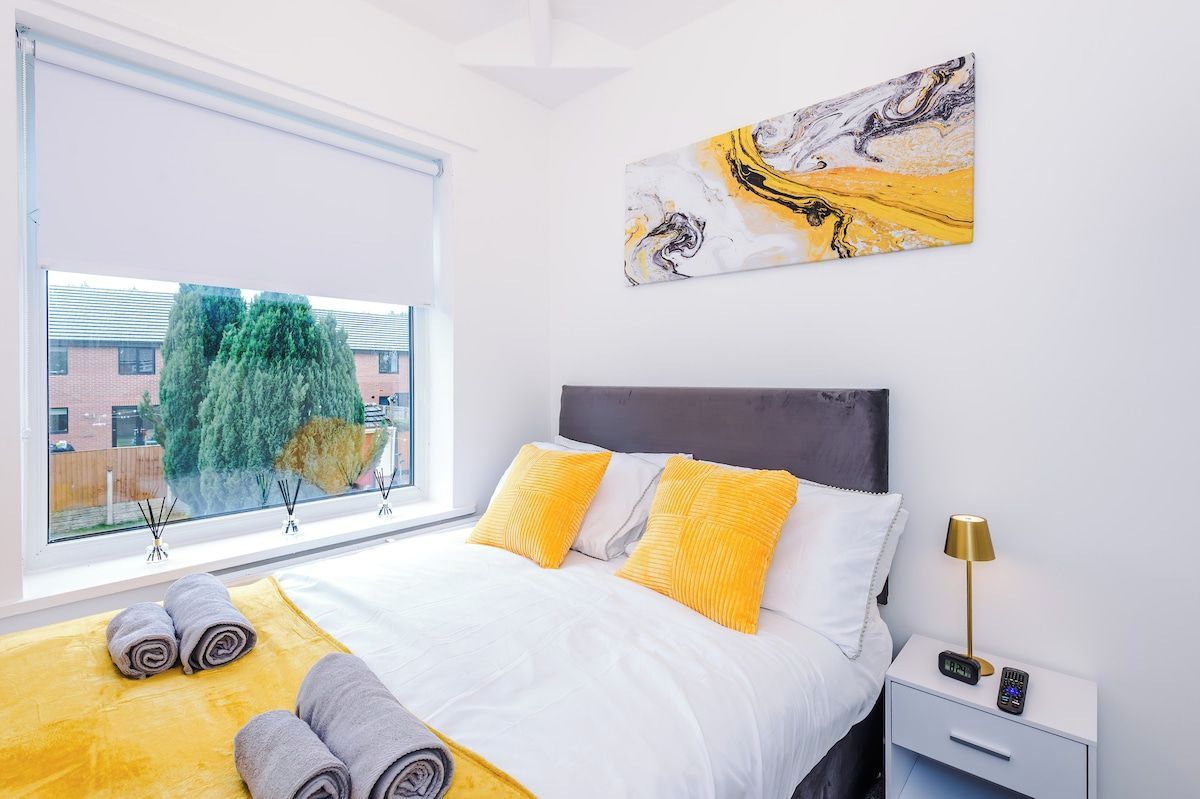Una moderna camera d'hotel con un letto grande, una finestra vista alberi, un quadro al muro e un comodino con una lampada.