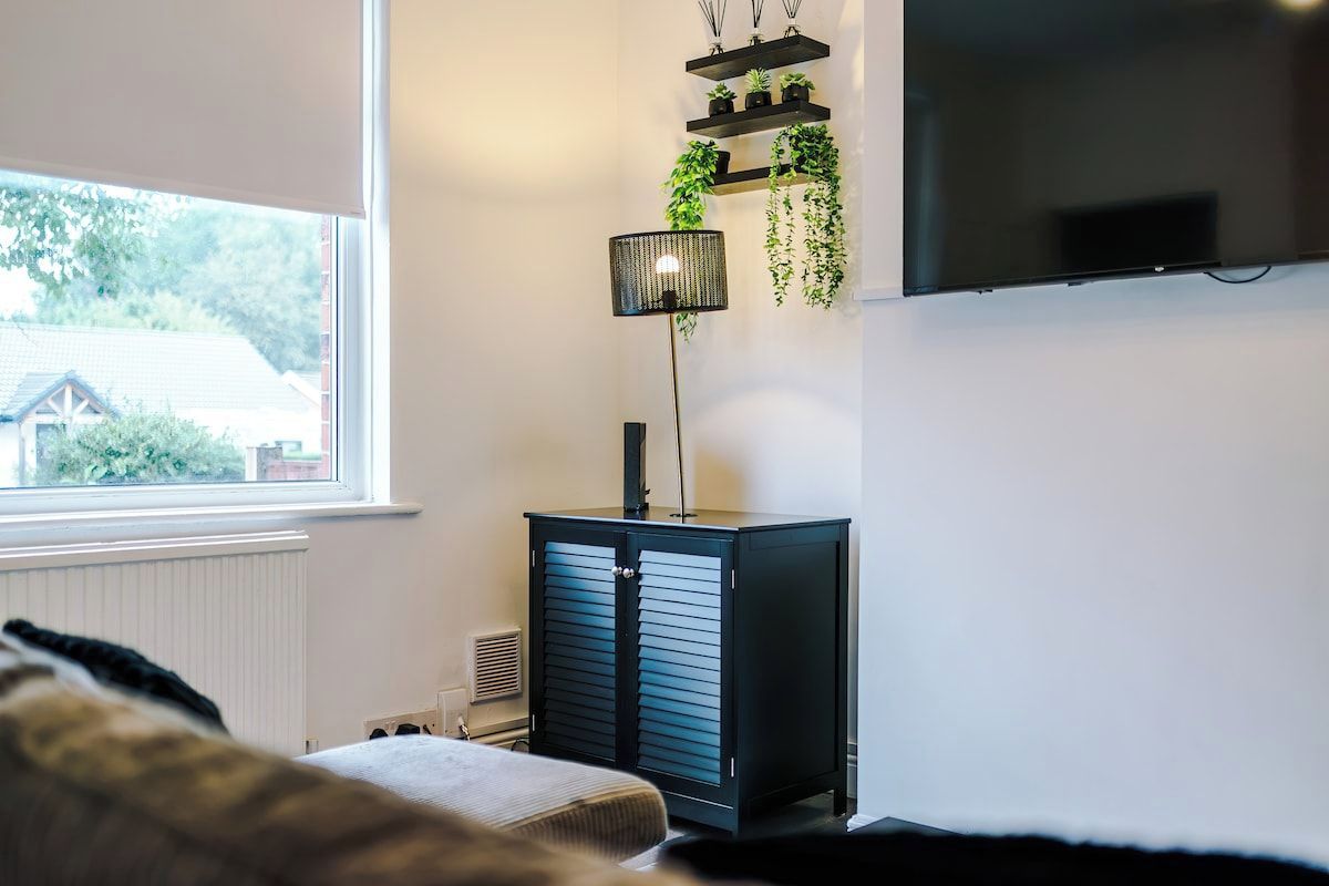 A room with a bed, a TV mounted on the wall, a radiator, and a window with a view of a house and trees.