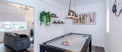 Una mesa de billar en una habitación con un sofá, una planta colgante y un cuadro en la pared.