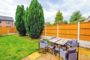 A patio with a table and chairs, surrounded by a wooden fence and greenery. - Stylish 3-Bed Home in Leigh | Garden & Parking (Greater Manchester)