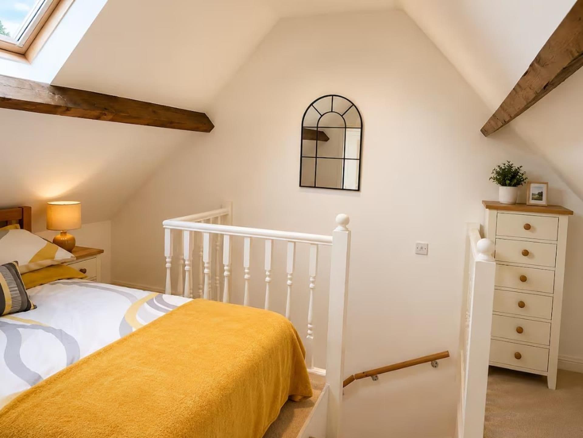 Une chambre avec un plafond en pente, un lit recouvert d’une couverture jaune, une table de chevet avec une lampe et une commode avec des tiroirs.