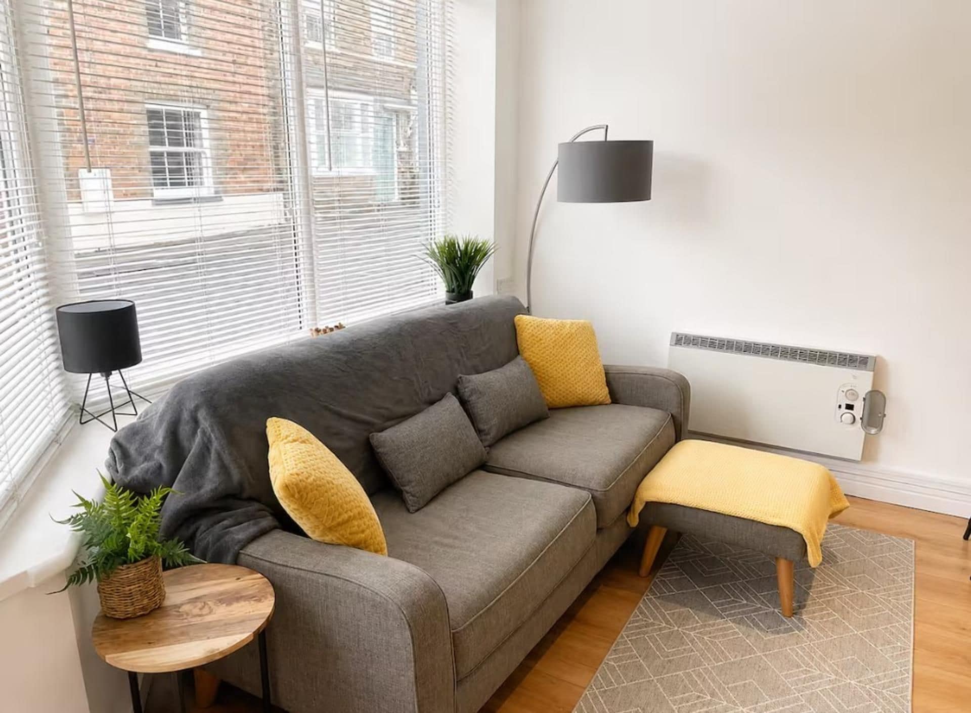 A modern living room with a grey sofa, yellow cushions, a small side table, a floor lamp, and a potted plant.