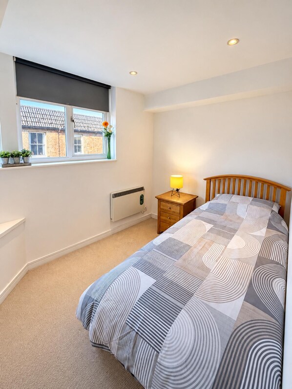 A bedroom with a bed, a bedside table with a lamp, a radiator, a window with a view, and a small shelf with plants.