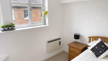 A bedroom with a window, radiator, bedside table, lamp, and a bed with a pillow.