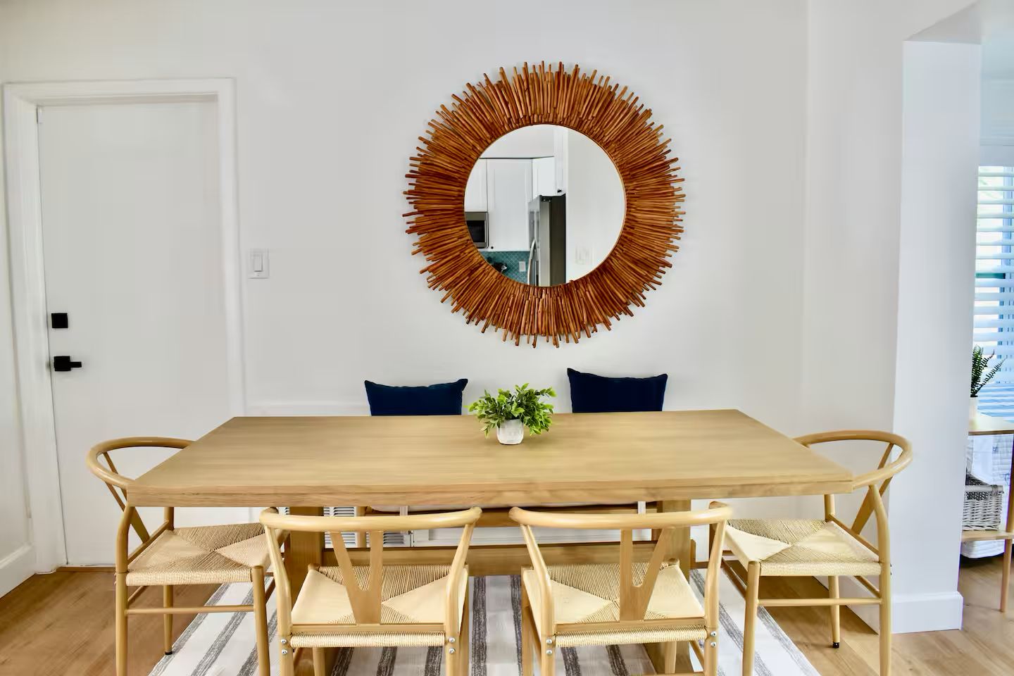 Une salle à manger avec une table en bois, six chaises, un miroir doté d’un cadre en forme de soleil et deux coussins foncés.