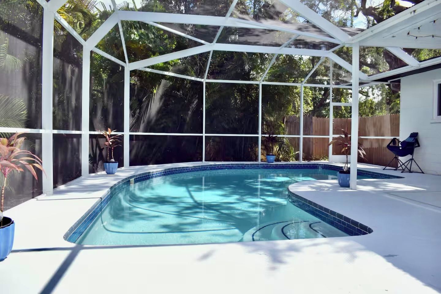 A covered swimming pool with a white roof and glass walls, surrounded by potted plants and a wooden fence.