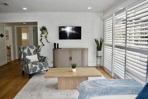 Ein Wohnzimmer mit einer Couch, einem Couchtisch, einem an der Wand befestigten Fernseher und einem Fenster mit Jalousien.