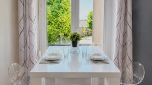 Et spiseområde med et hvidt bord, to stole, en potteplante og et vindue med udsigt til grønne områder.