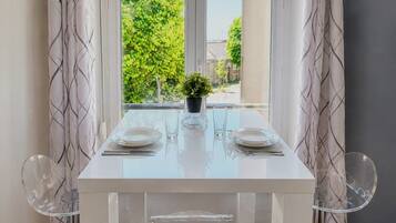 Un coin repas avec une table blanche, deux chaises, une plante en pot et une fenêtre donnant sur un paysage verdoyant.