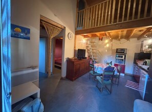 A cozy living room with a dining area, wooden beams, a staircase, and a kitchen with appliances. - Maison en ossature bois à Vensac Océan (Vensac)