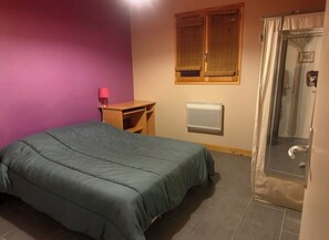 A room with a bed, a desk, a window with blinds, a radiator, and a shower area. - Maison en ossature bois à Vensac Océan (Vensac)