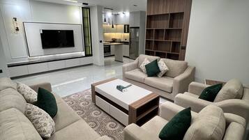 A modern living room with a flat-screen TV, beige sofas, a coffee table, and a kitchen in the background.