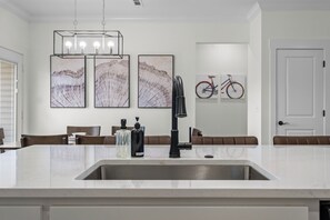 A modern kitchen with a white marble countertop, a stainless steel sink, and a black faucet. There are three framed artworks on the wall, a bicycle, and a door in the background. - 3-bedroom house in charming Bella Vista with AC for relaxing stays (Bella Vista)