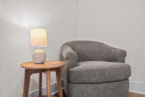A corner of a room with a grey upholstered armchair and a wooden side table with a lamp.
