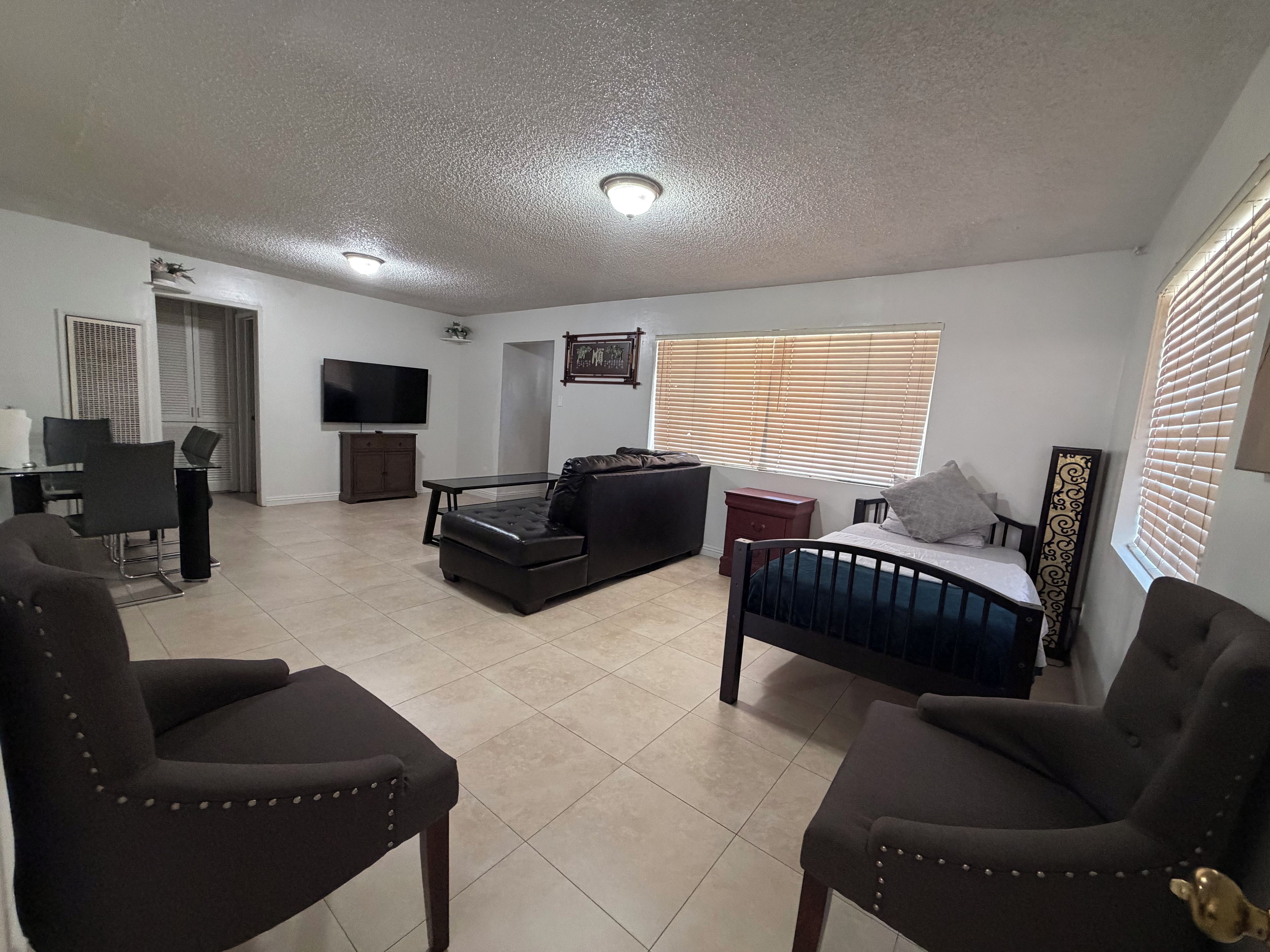 A living room with a sofa, two armchairs, a TV, a dining area with chairs, and a window with blinds.