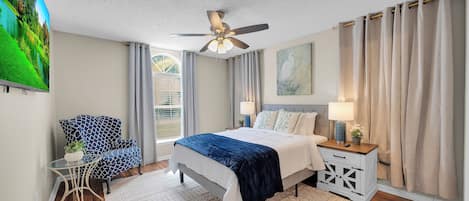 Une chambre à coucher comprenant un lit, une chaise, une table de chevet, une lampe, un tableau et un ventilateur de plafond.