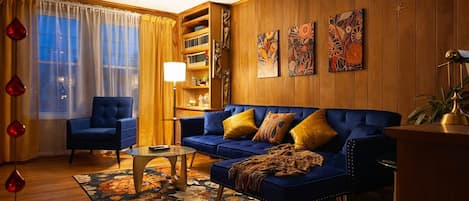 A living room with a blue sofa, a blue armchair, a coffee table, and a bookshelf against a wooden paneled wall.