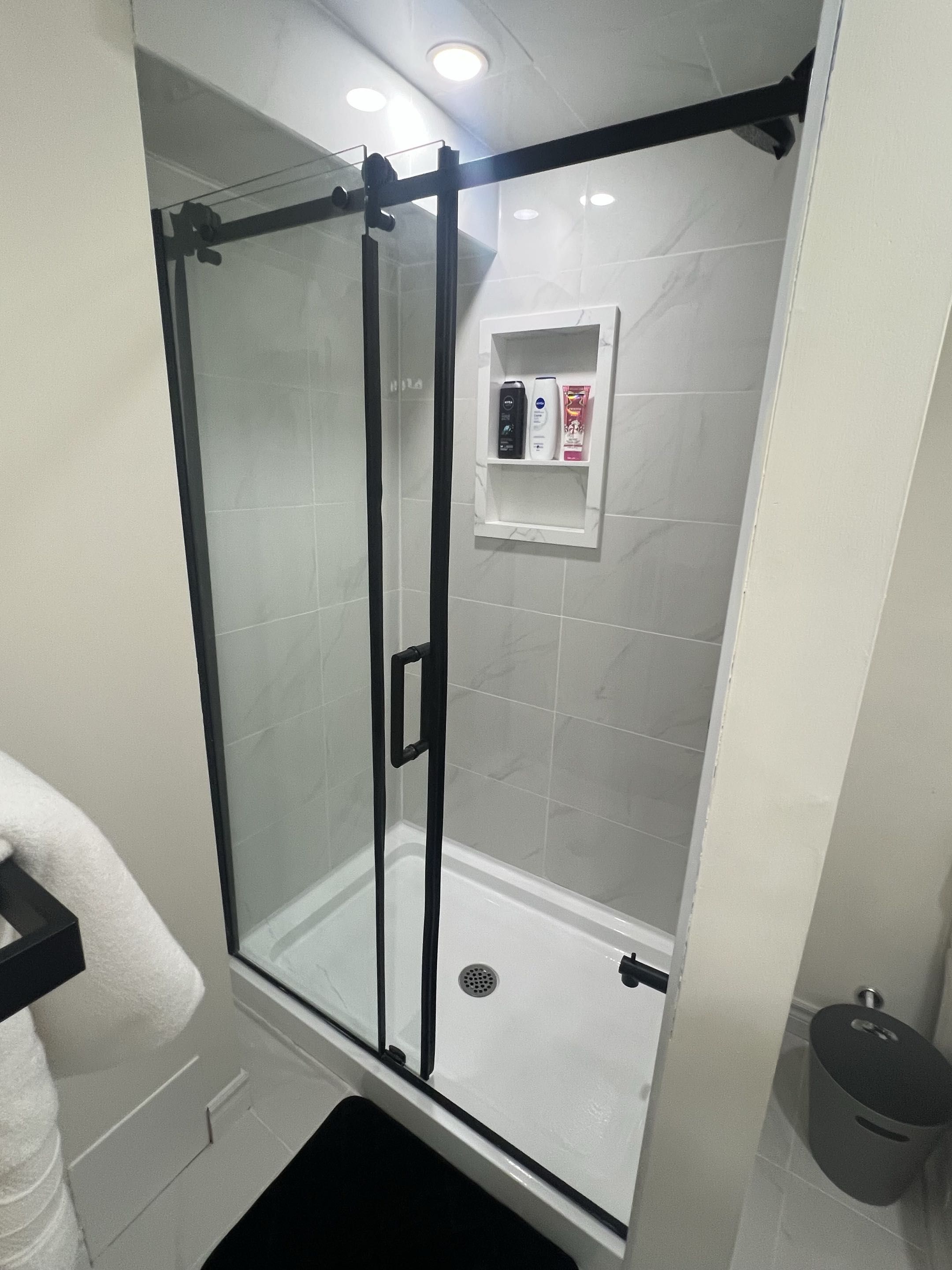 Ducha envuelta en vidrio con panel de control, paredes de azulejos blancos y un tapete de piso negro.