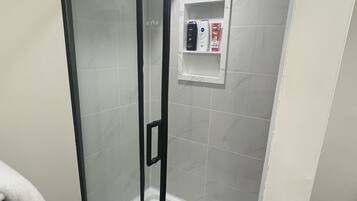 Ducha envuelta en vidrio con panel de control, paredes de azulejos blancos y un tapete de piso negro.