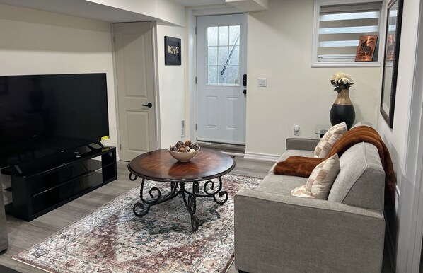 Een woonkamer met een flatscreen televisie, een bijzettafeltje met decoratieve objecten, een bank met kussens en een kamerkleed met een patroon.