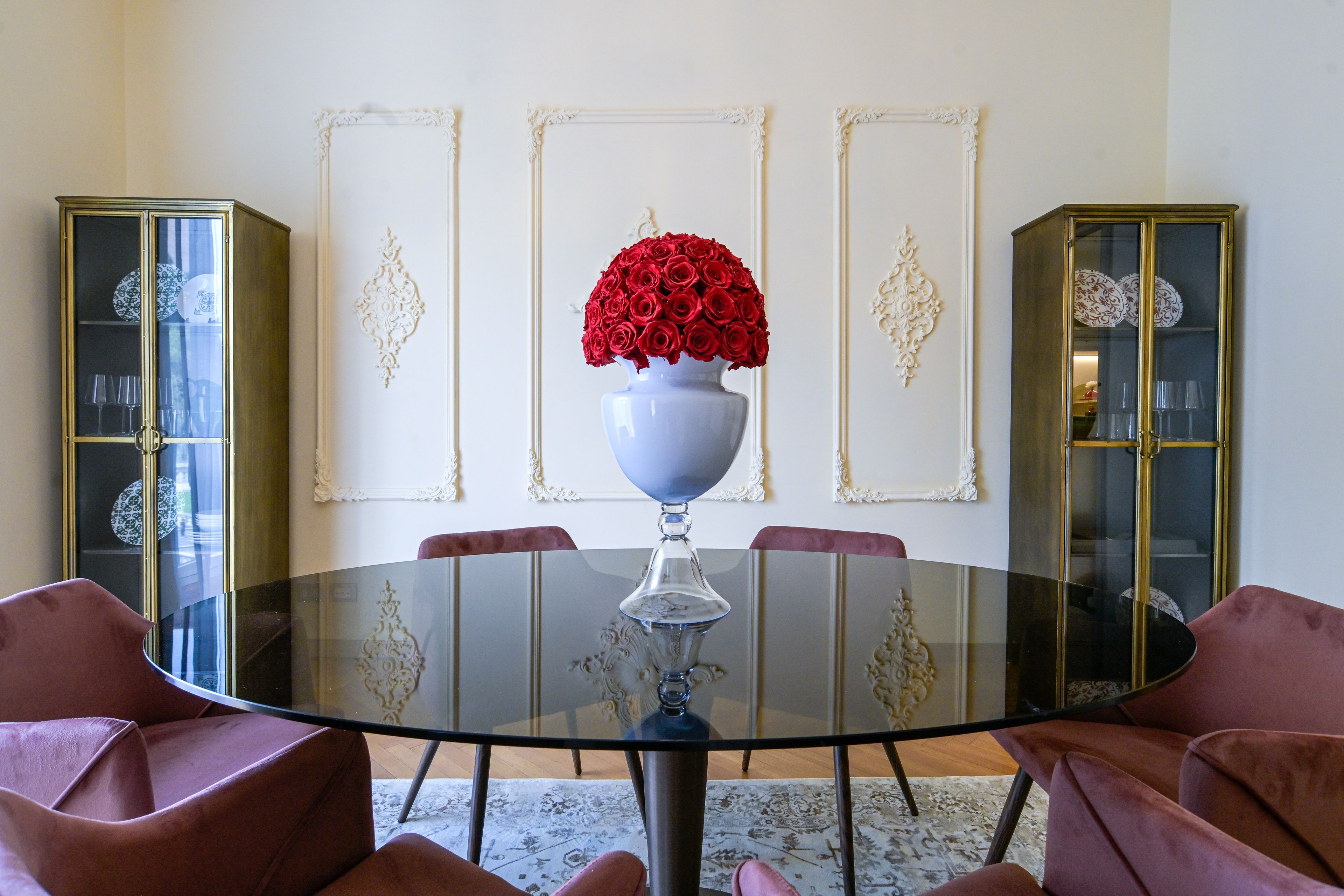 Ein Essbereich mit einem runden Glastisch, roten Rosen in einer weißen Vase und zwei Anrichteschränken mit Glastüren.