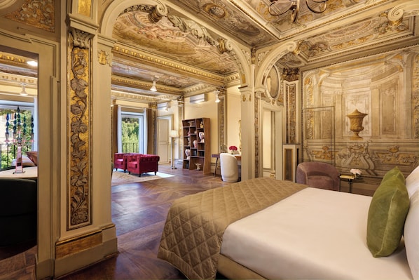 Una lujosa habitación de hotel con una cama grande, un sofá rojo y una estantería. La habitación tiene decoraciones ornamentales en el techo y vistas a un jardín.