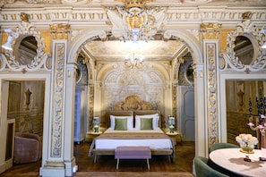Un dormitorio ornamentado con una cama grande, dos mesitas de noche, una araña y decoraciones elaboradas en las paredes.