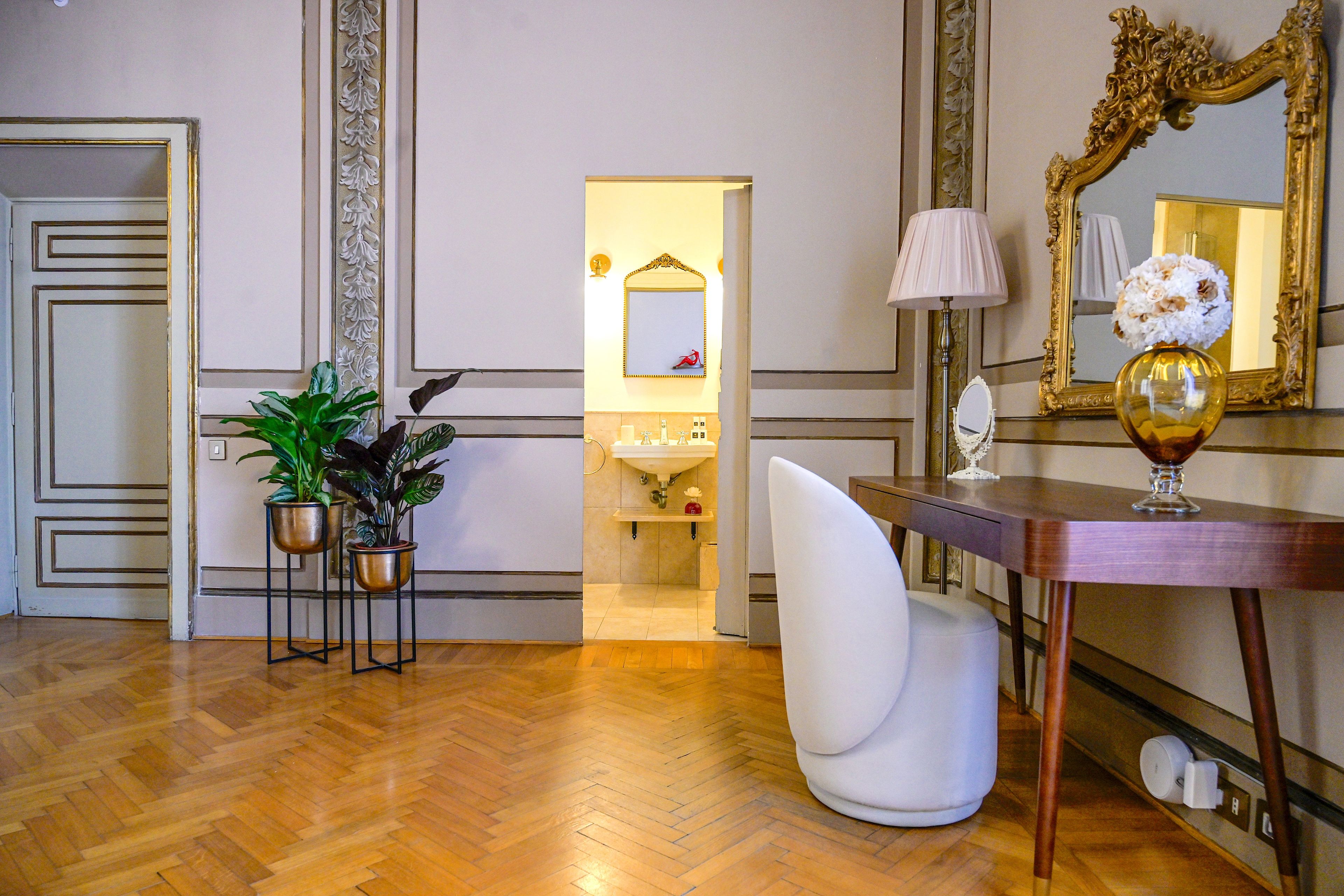 A room with a wooden floor, a white chair, a desk with a lamp, a mirror, and two potted plants.