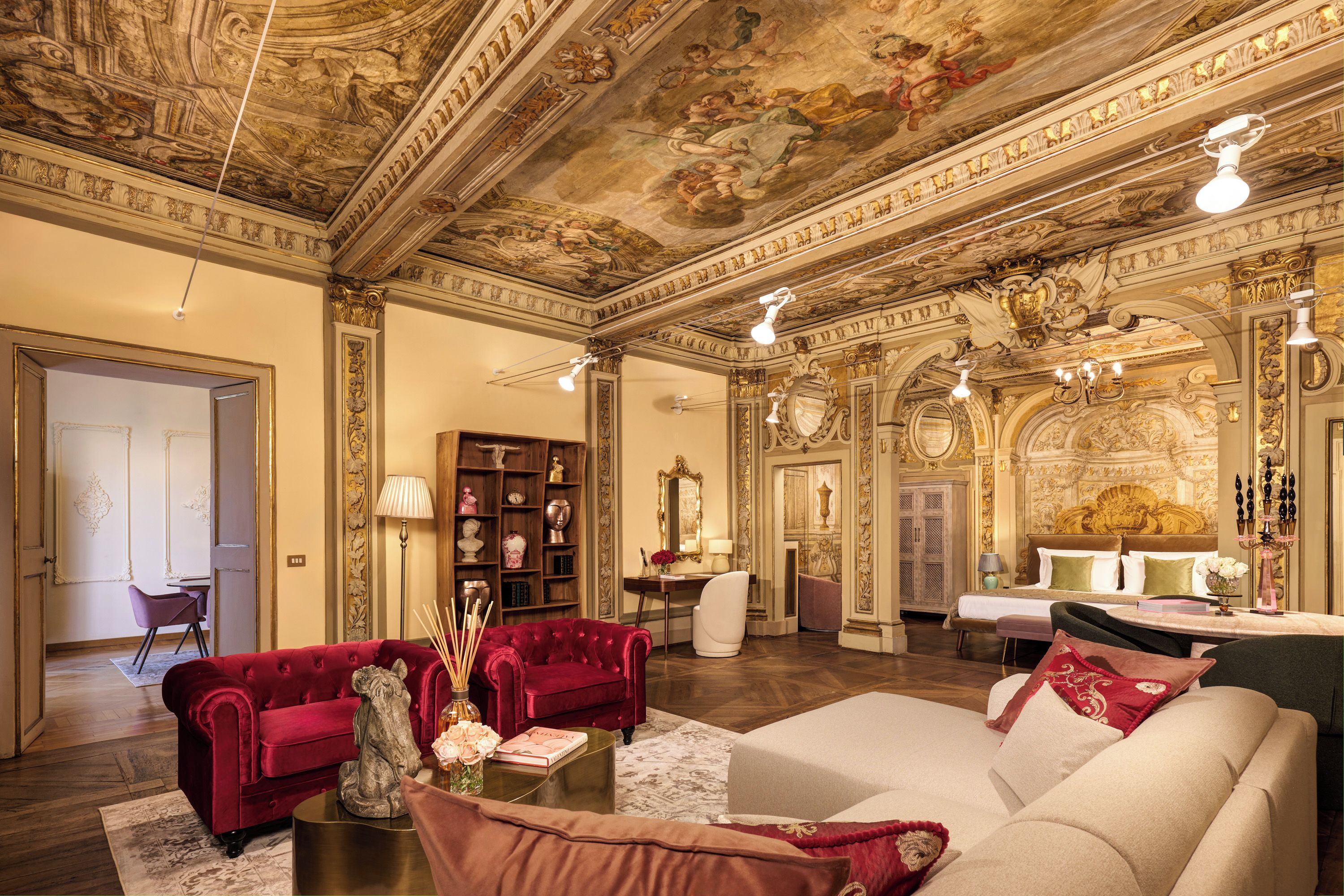 Una habitación lujosa con frescos ornamentales en el techo, un sofá rojo con tapicería suave, un sofá blanco y una cama clásica.