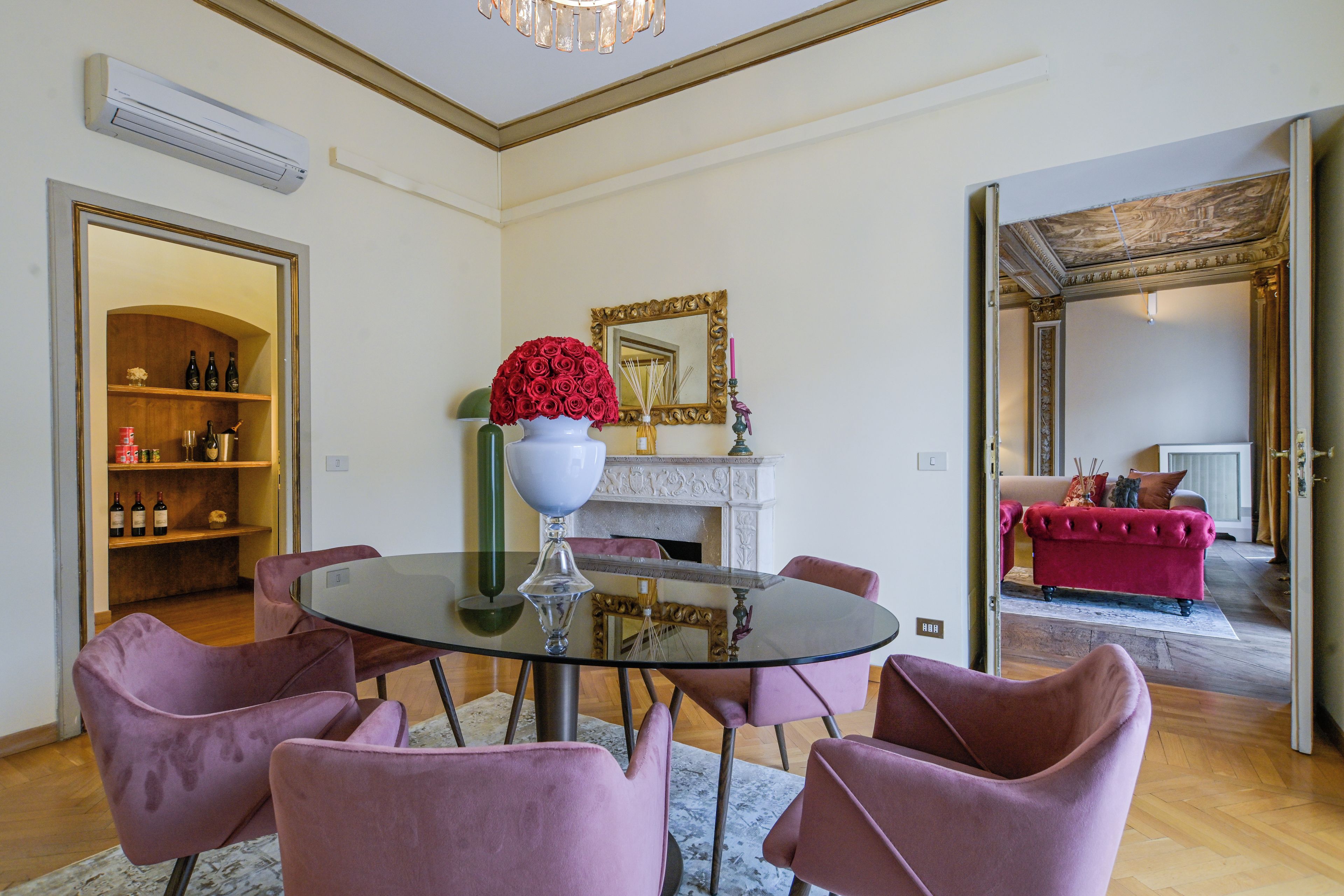 A room with a round glass table, pink chairs, a white vase with red flowers, a mirror with a gold frame, and a red sofa in the background.