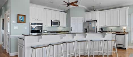 Una cocina con armarios blancos, una isla central con taburetes, un ventilador de techo y un microondas.