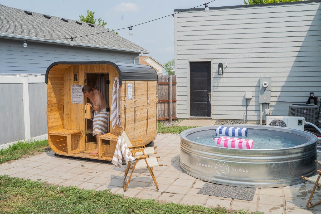 Un sauna avec une façade en bois et une personne à l’intérieur, un jacuzzi avec des coussins à rayures, et une maison en arrière-plan.
