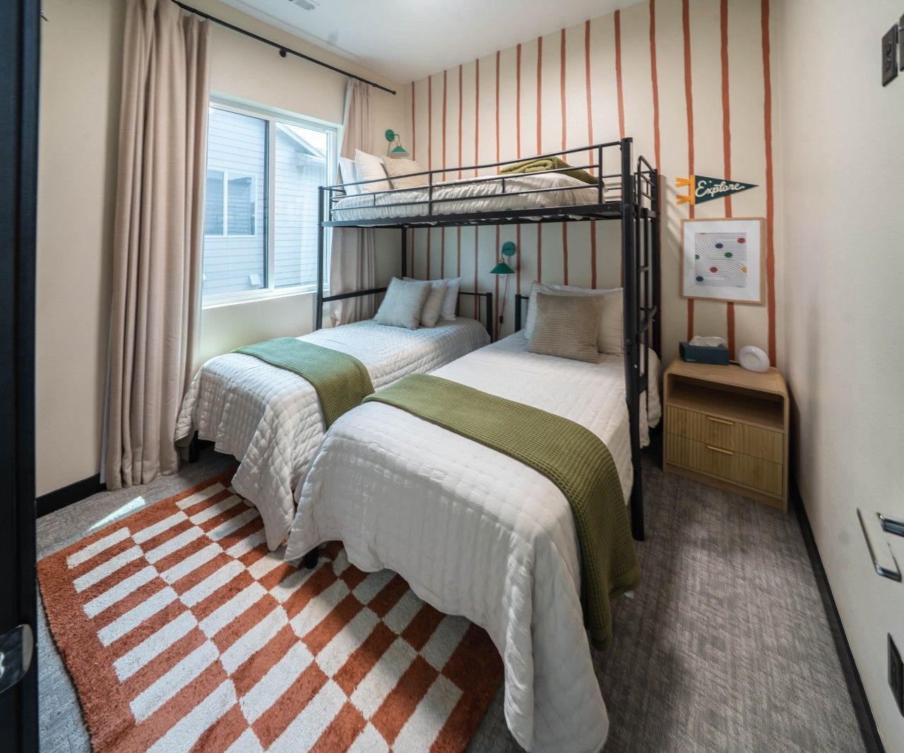 Un dormitorio con dos camas, una litera, una ventana con cortinas, una alfombra, una mesita de noche y una pared con papel tapiz a rayas.