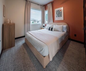 A hotel room with a large bed, a wooden nightstand, a window with curtains, and a wall-mounted artwork.