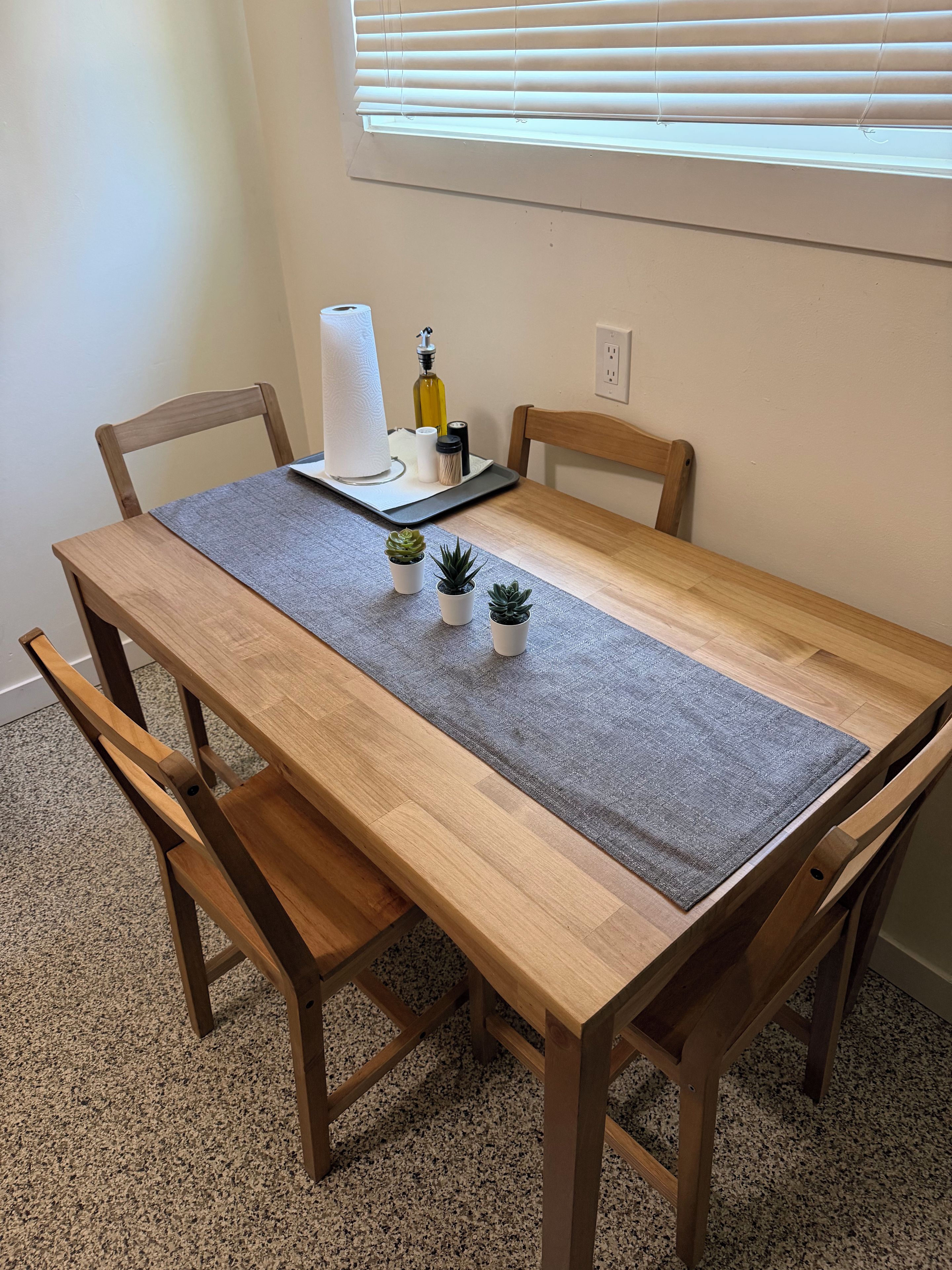 A wooden dining table with a grey placemat, four wooden chairs, and three small potted plants.