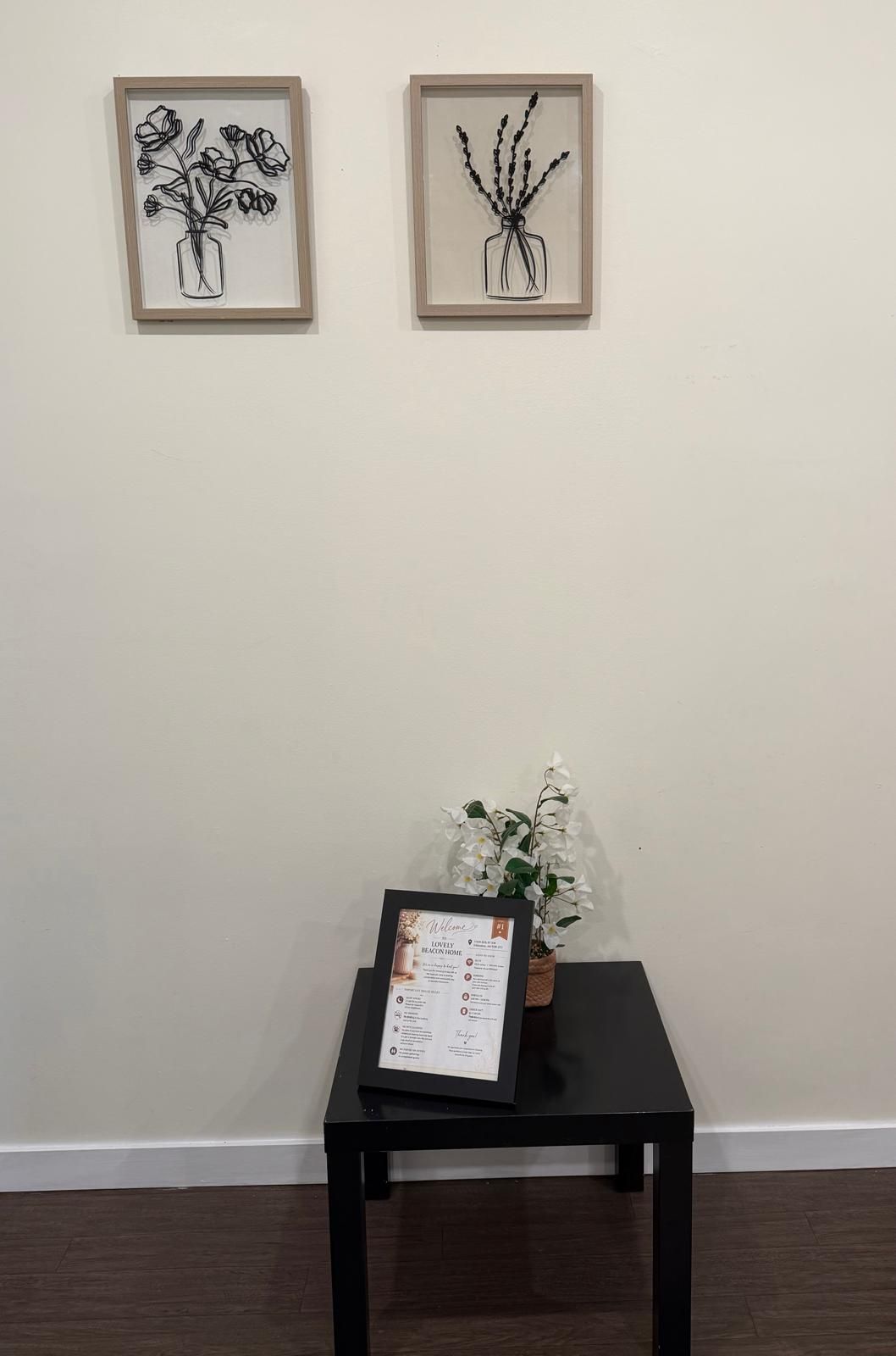 A framed artwork of a vase with flowers on a wall, a small table with a potted plant and a welcome sign, and a dark wooden floor.