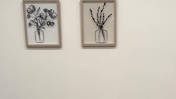 A framed artwork of a vase with flowers on a wall, a small table with a potted plant and a welcome sign, and a dark wooden floor.