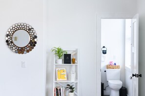Ein Badezimmer mit Toilette, einem weißen Regal mit Büchern und einer Topfpflanze sowie einem Spiegel mit runder Umrandung.