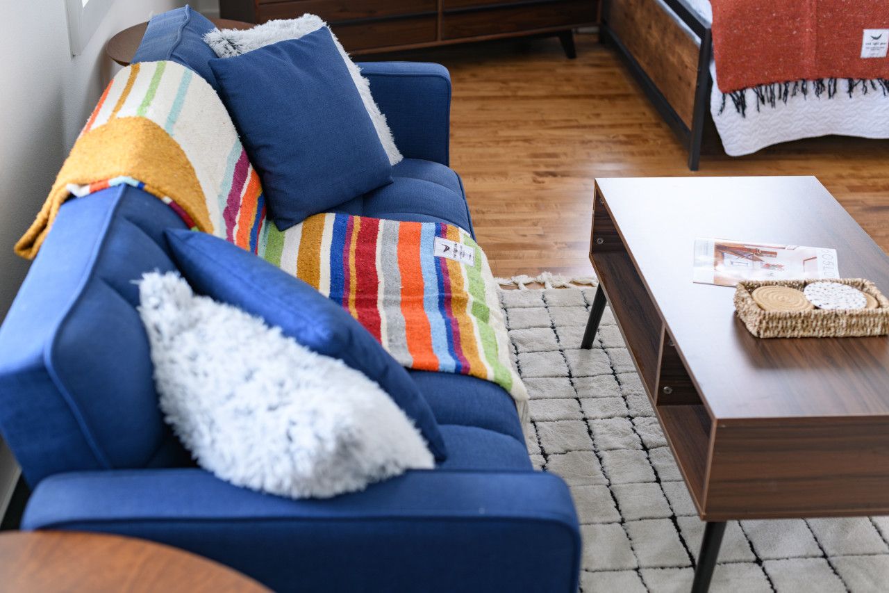 A blue sofa with colorful pillows and a white fluffy pillow, a wooden coffee table with a woven basket, and a patterned rug on a wooden floor.