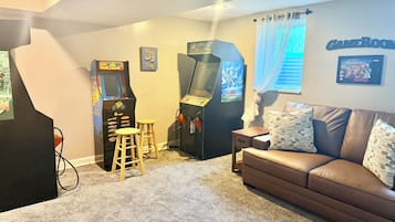 Una stanza con un divano in pelle marrone, due cabinati arcade, uno sgabello e un cartello fissato al muro con la scritta 'GAMEROOM'.