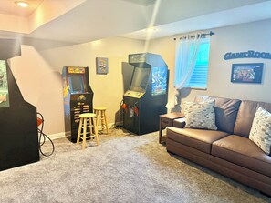 A room with a brown leather couch, two arcade machines, a small stool, and a wall-mounted sign that reads 'GAMEROOM'.