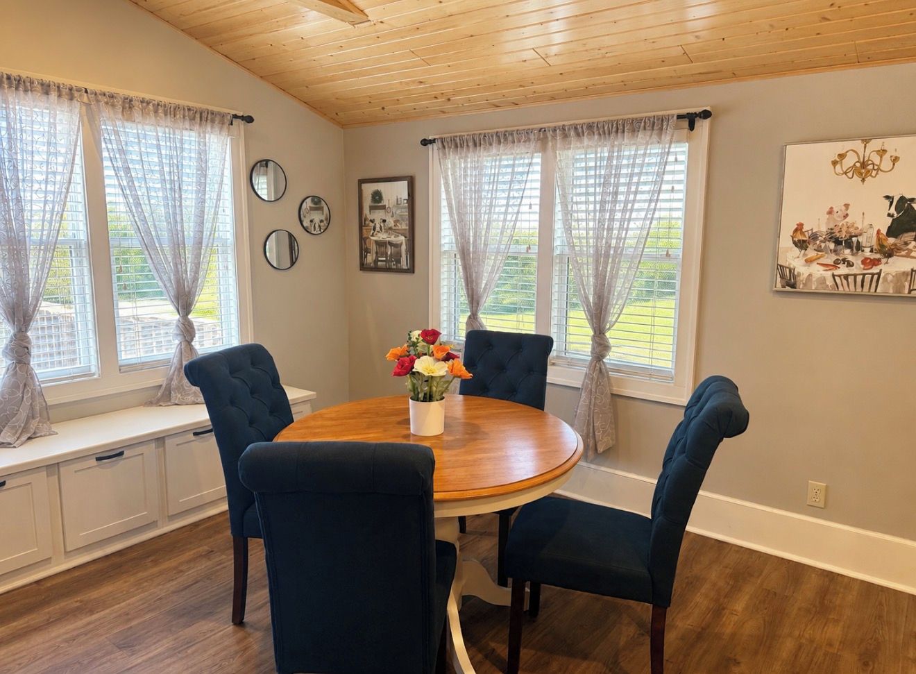 A dining area with a round table, four chairs, a vase of flowers, and curtains on windows.
